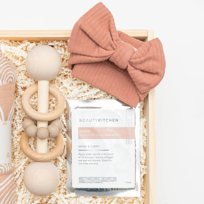Celebrate the arrival of a new baby with this modern, elegant and practical gift box. Close up of Organic Cotton Headband, Natural Wood Rattle, Naked Collagen Eye Gels,